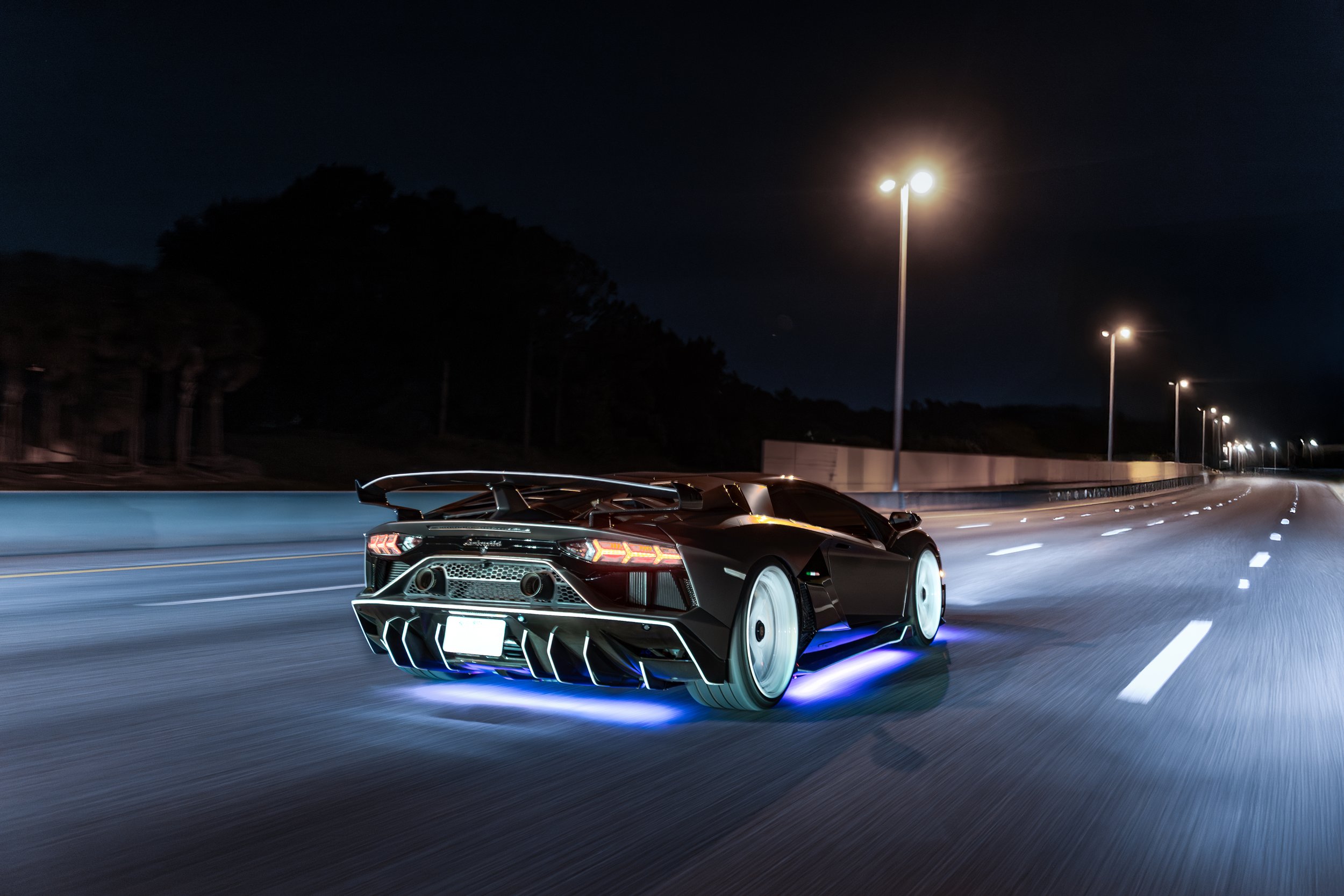 A black Lamborghini SVJ with illuminated underglow driving on an empty highway at night owned by MyExotics