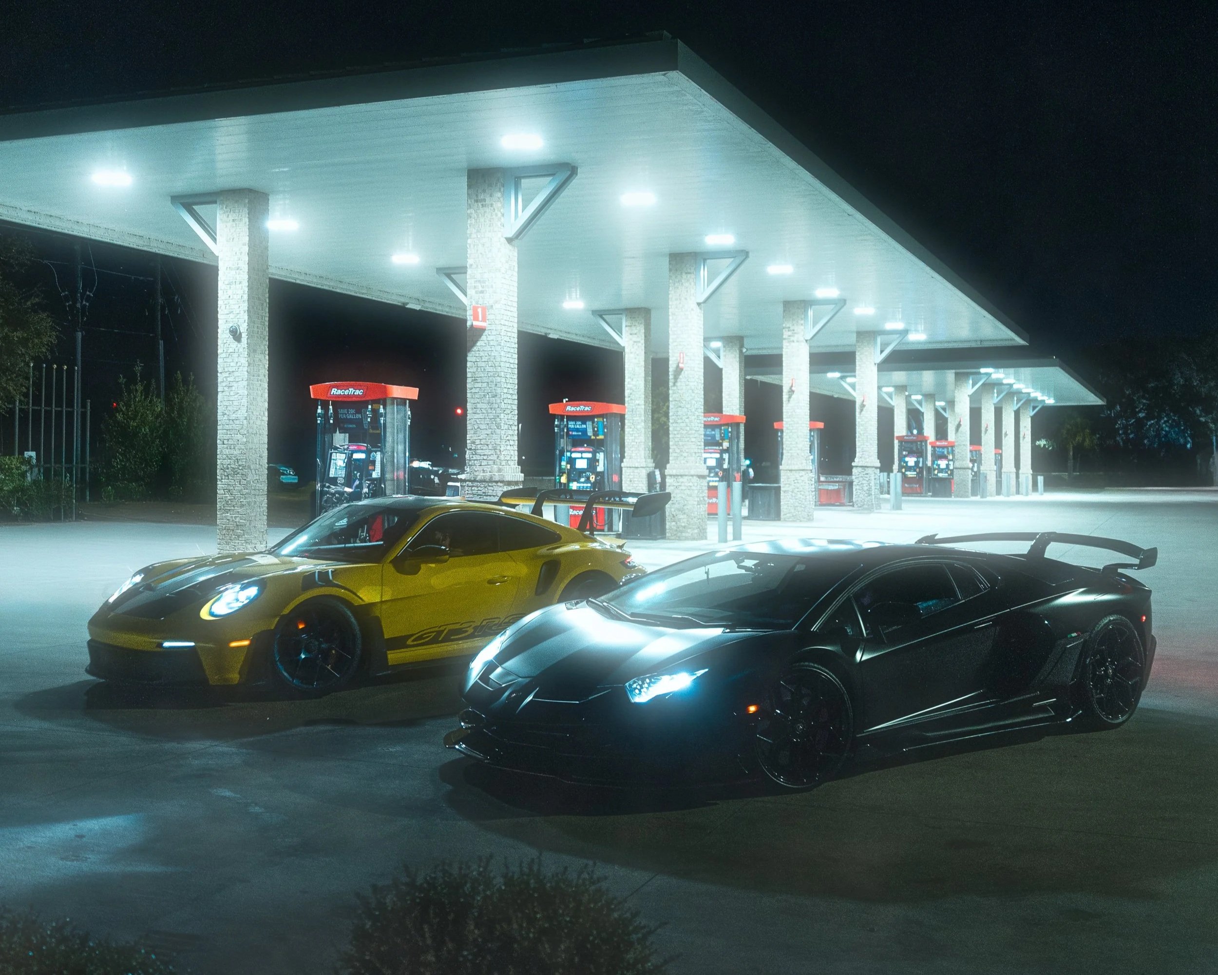Nighttime scene at a gas station with two luxury sports cars, a yellow Porsche and a black Lamborghini SVJ, parked in front.