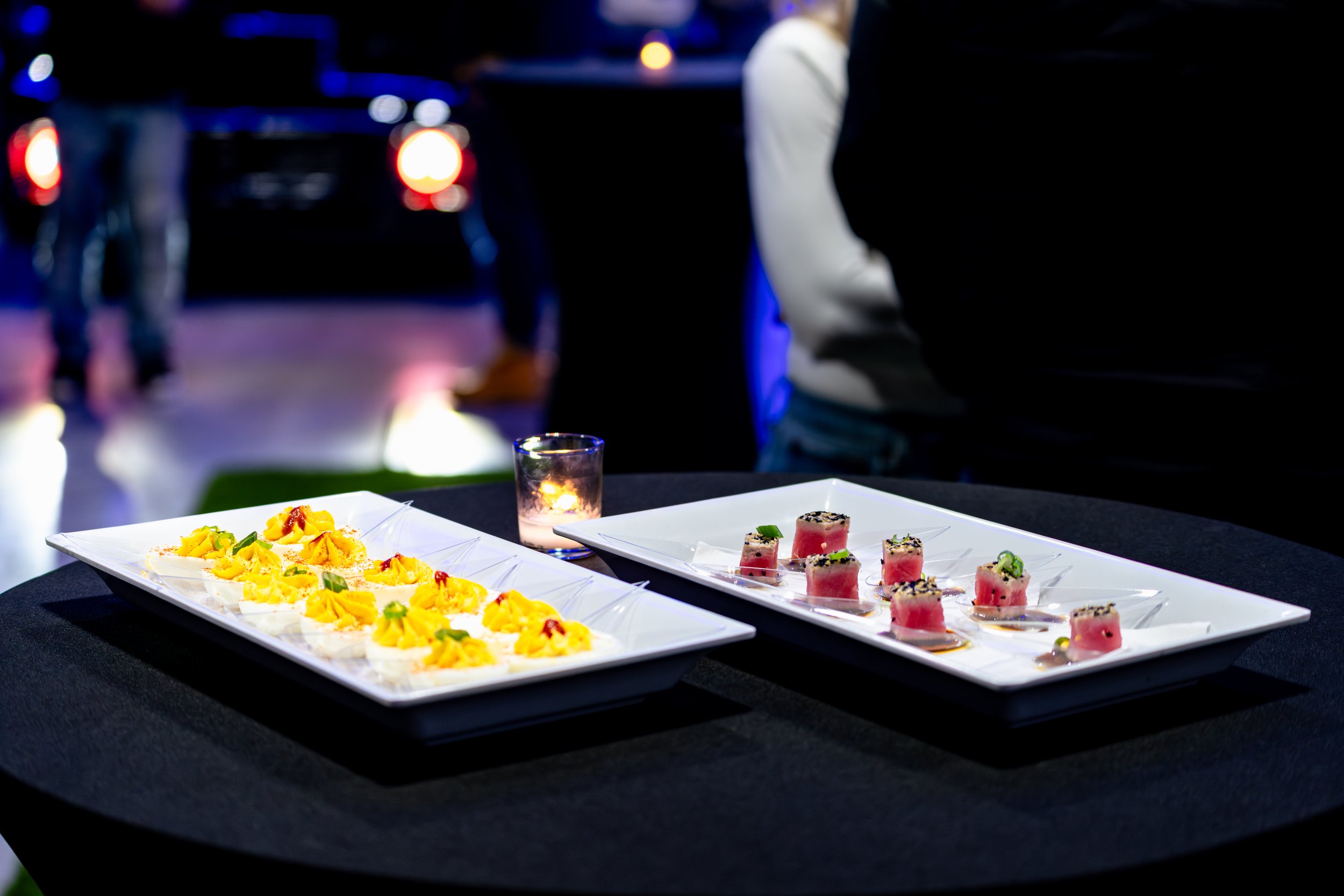 Two rectangular white plates with small culinary dishes on a black table, surrounded by a candle in a small glass holder. The background shows blurred people and a dimly lit environment, an event at Rochester Car Club