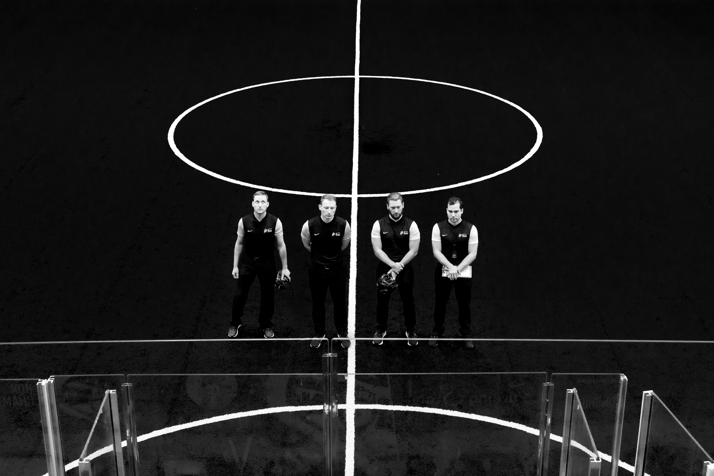 Four referees standing on an empty indoor basketball court with a black surface and white lines, seen from above at the World Box Lacrosse Tournament in Utica NY