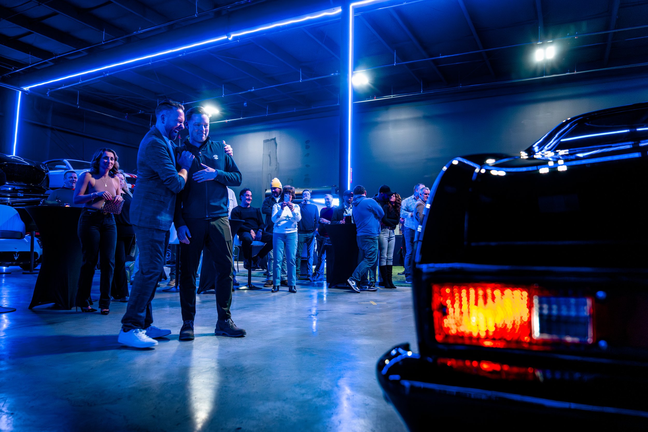 People gathered in a showroom with neon blue lighting and luxury cars, some standing and talking, others taking photos, at a social event at the Car Club of Rochester