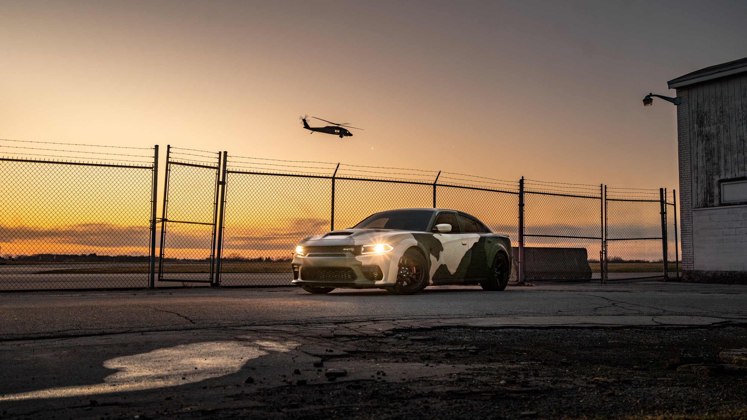 A white and black camouflage patterned sports car parked on a cracked pavement in front of a chain-link fence during sunset, with a helicopter flying in the sky above near a building.