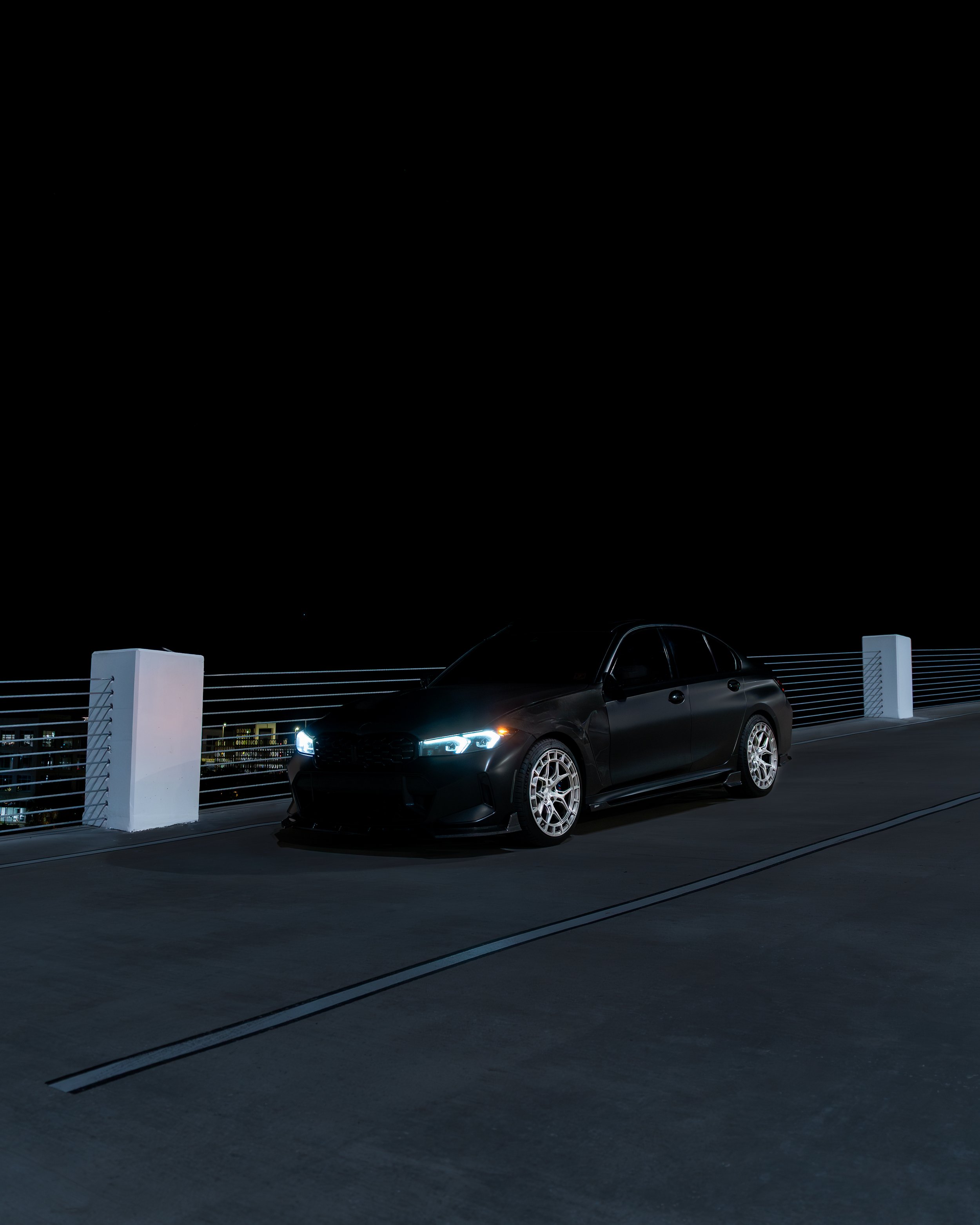 Black car with illuminated headlights parked on a rooftop at night, with city lights in the background.