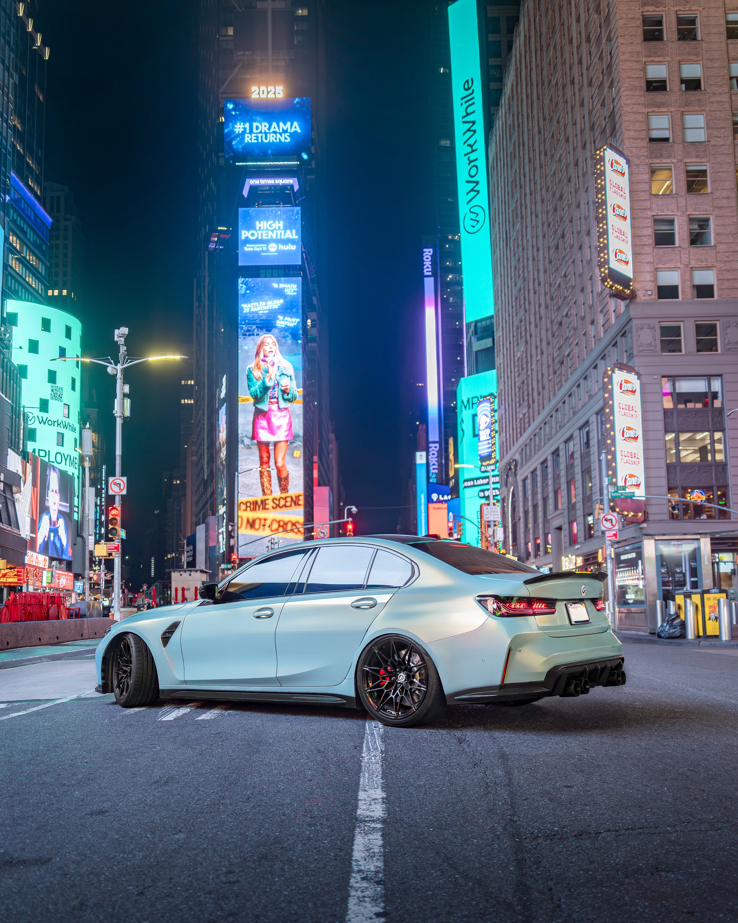 A white sports sedan parked in Times Square at night with bright digital billboards and signs illuminated in the background.