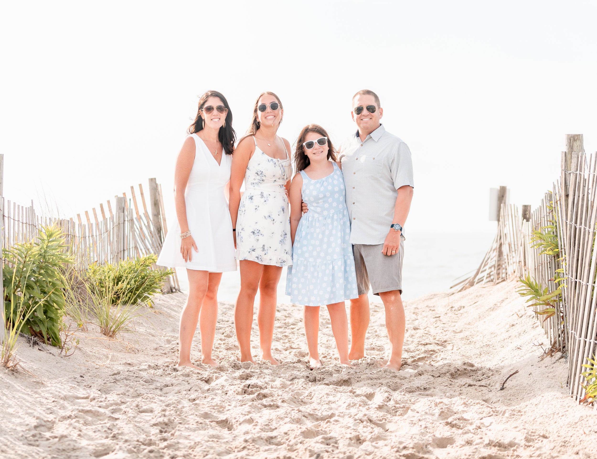 A family of four standing on a sandy beach pathway with fencing and greenery on either side, smiling at the camera.