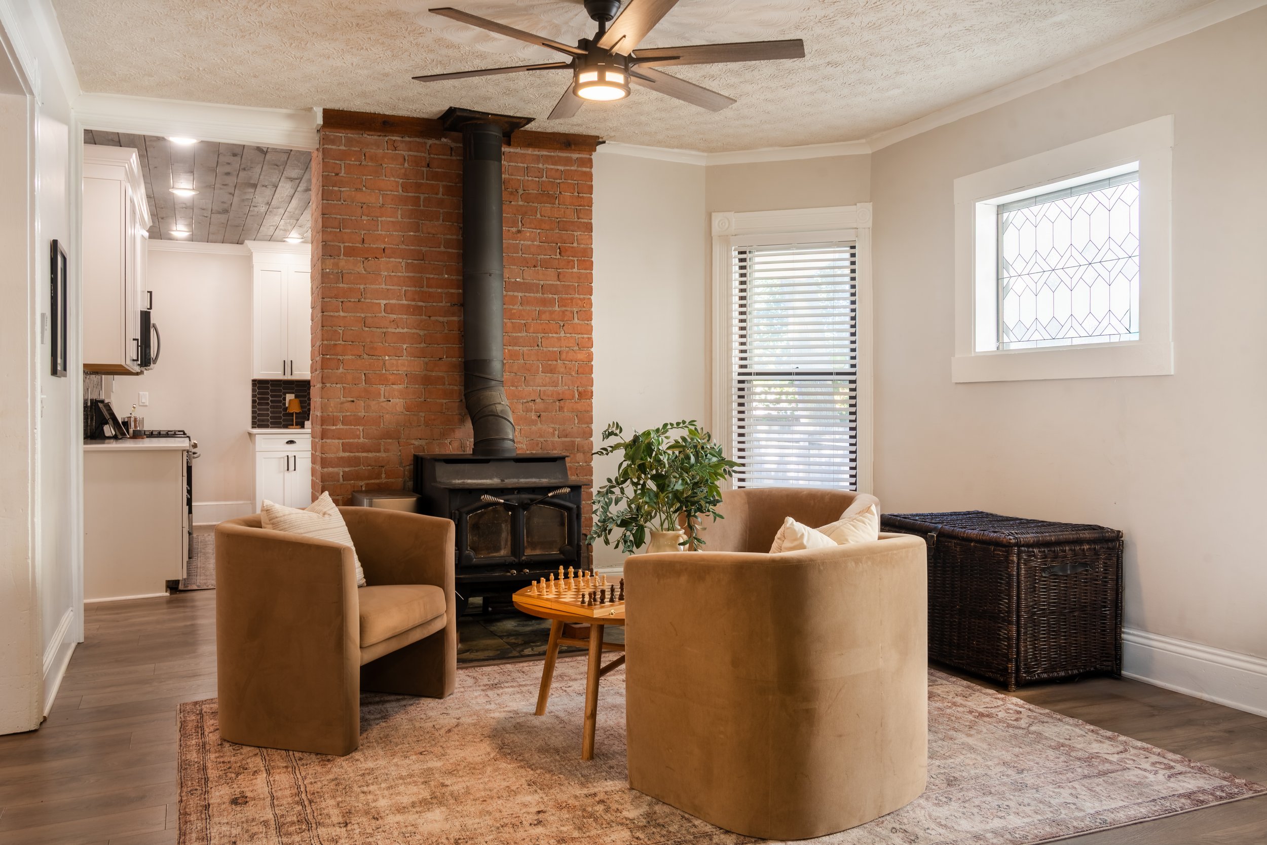 Living room with beige armchairs around a small coffee table in front of a black wood stove, with a brick chimney and a ceiling fan. There are windows with blinds, a plant, and a woven storage chest.