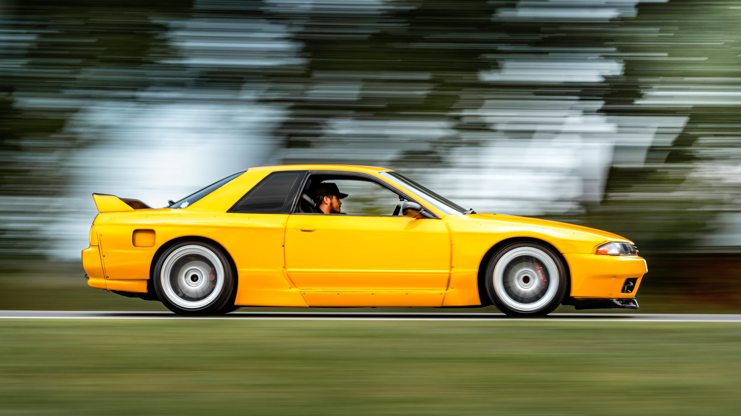 A yellow sports car in motion on a road with a blurred background, driven by a man with a black cap.