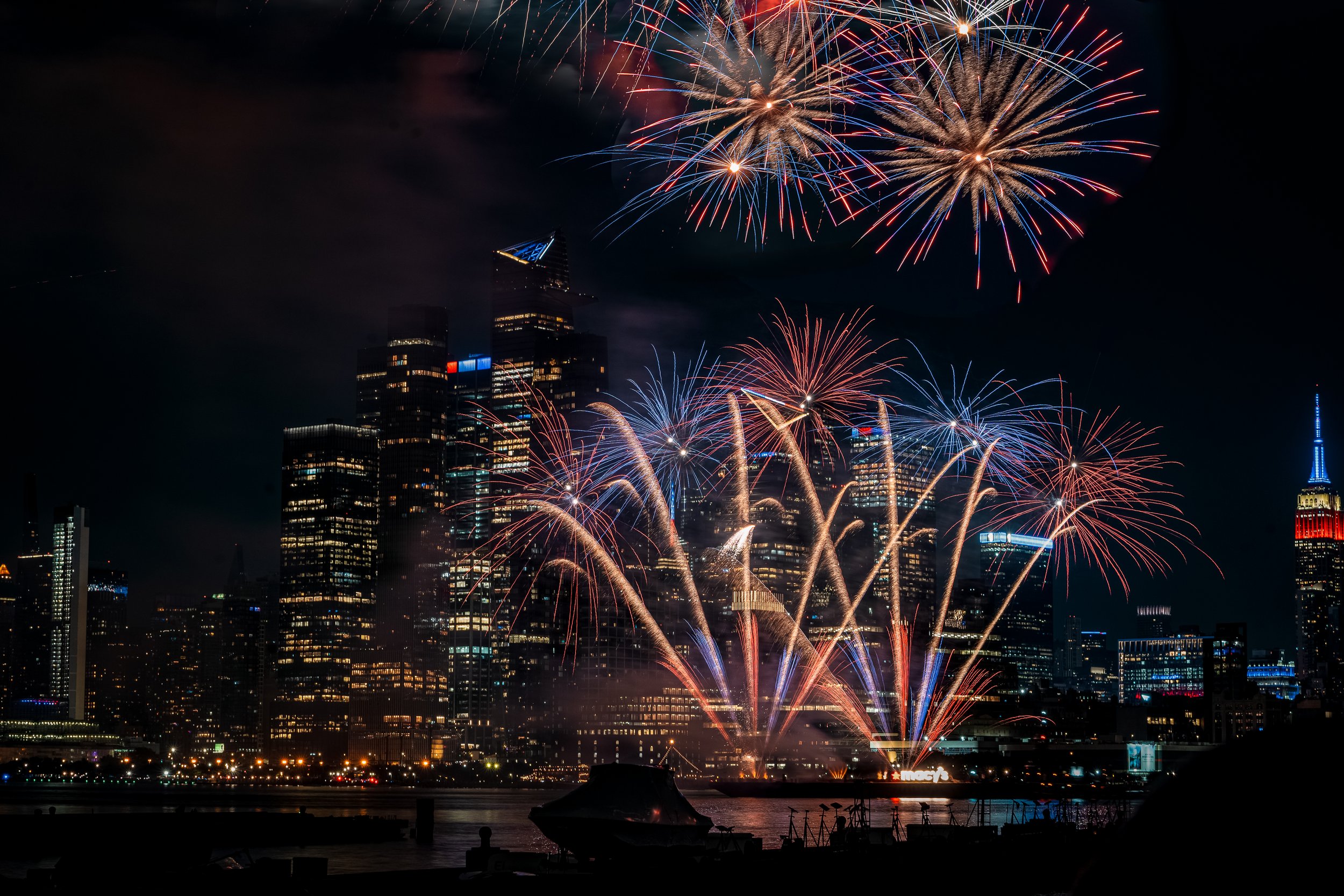 Fireworks display over a city skyline at night, with tall buildings illuminated and colorful fireworks bursting in the sky.