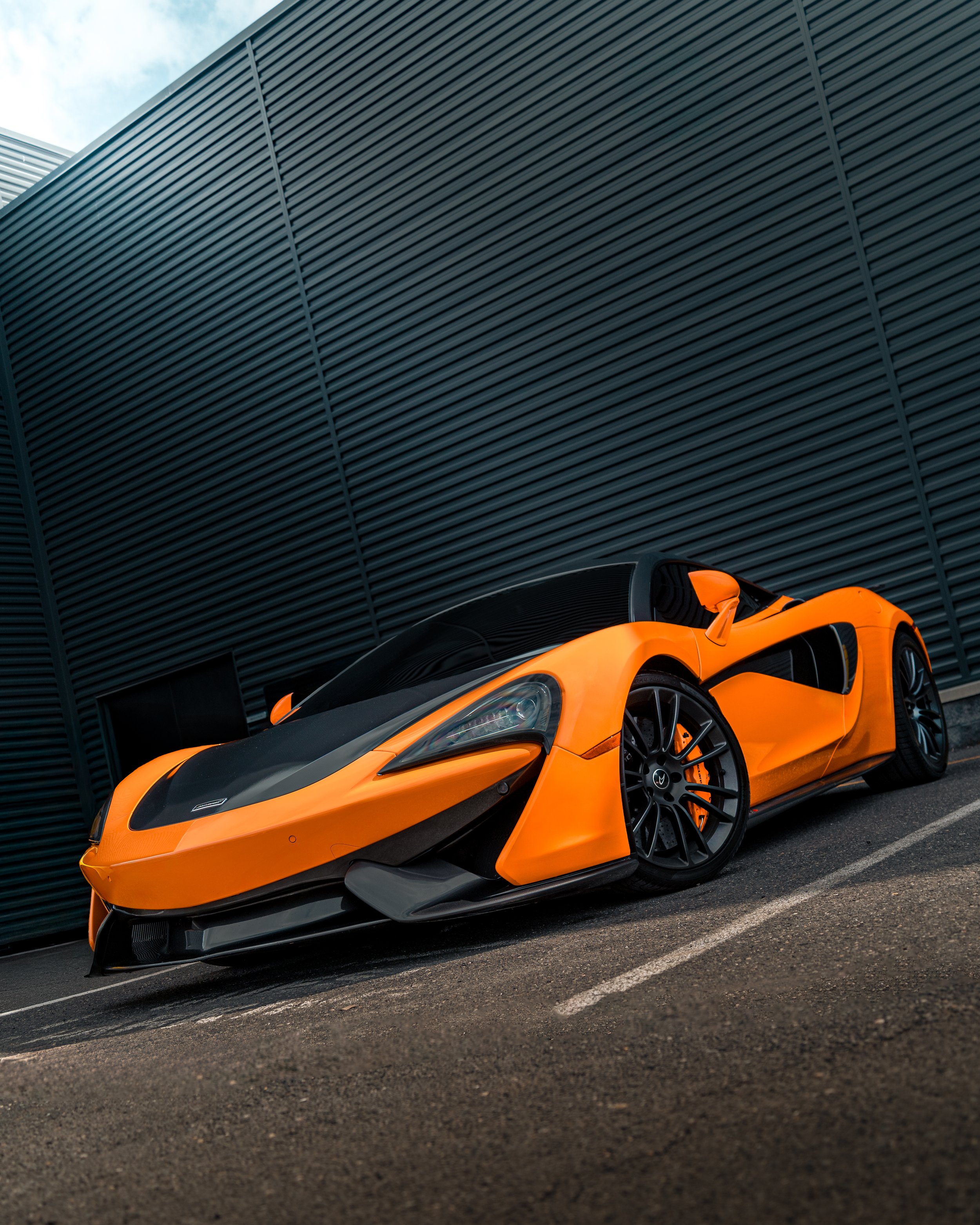 An Mclaren 570s car parked in front of a modern building with dark gray, vertically ribbed exterior walls.