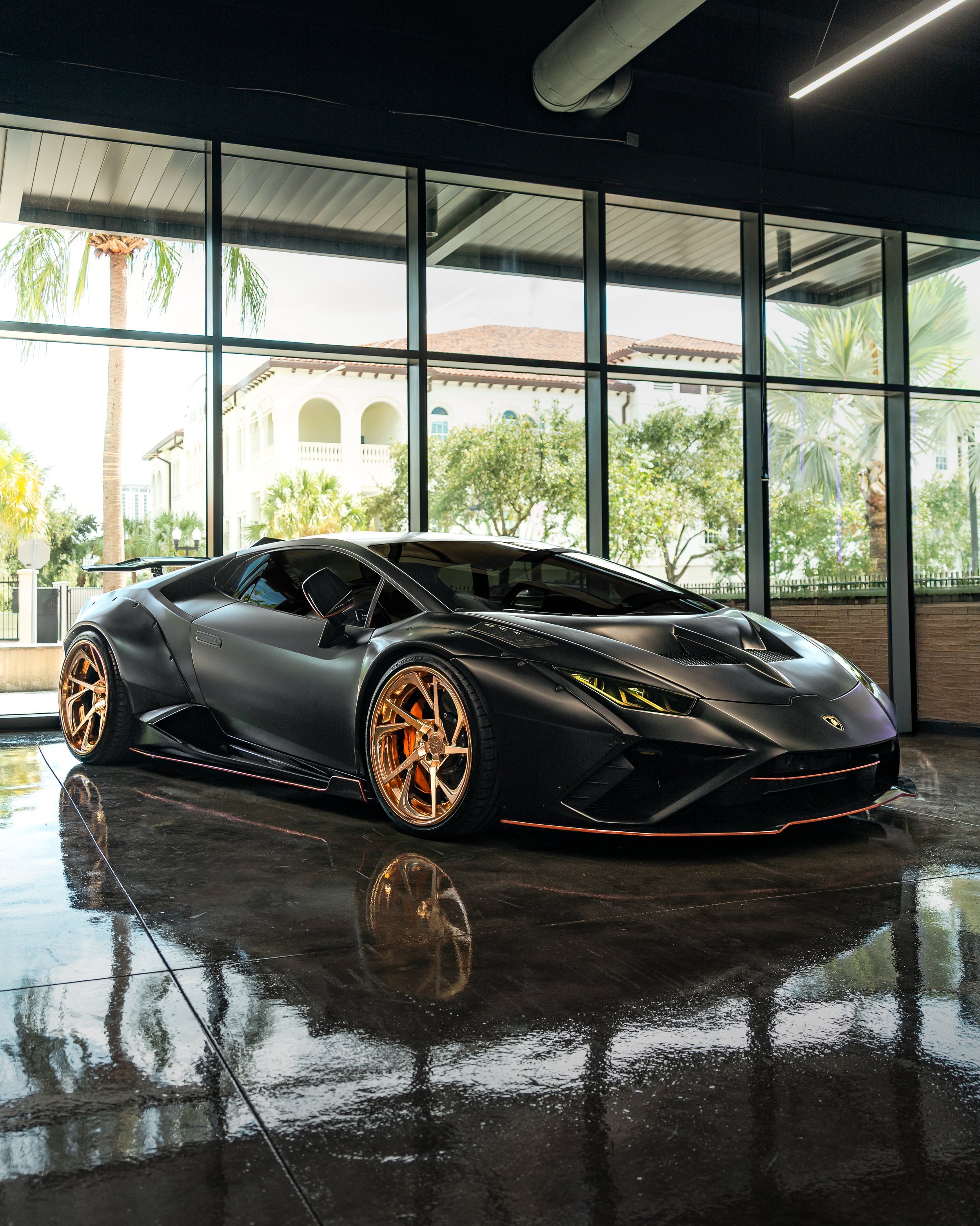 A black Liberty Walk widebody one of one Lamborghini Huracan Performante sports car with gold wheels parked indoors near large glass windows showing palm trees and residential buildings outside.