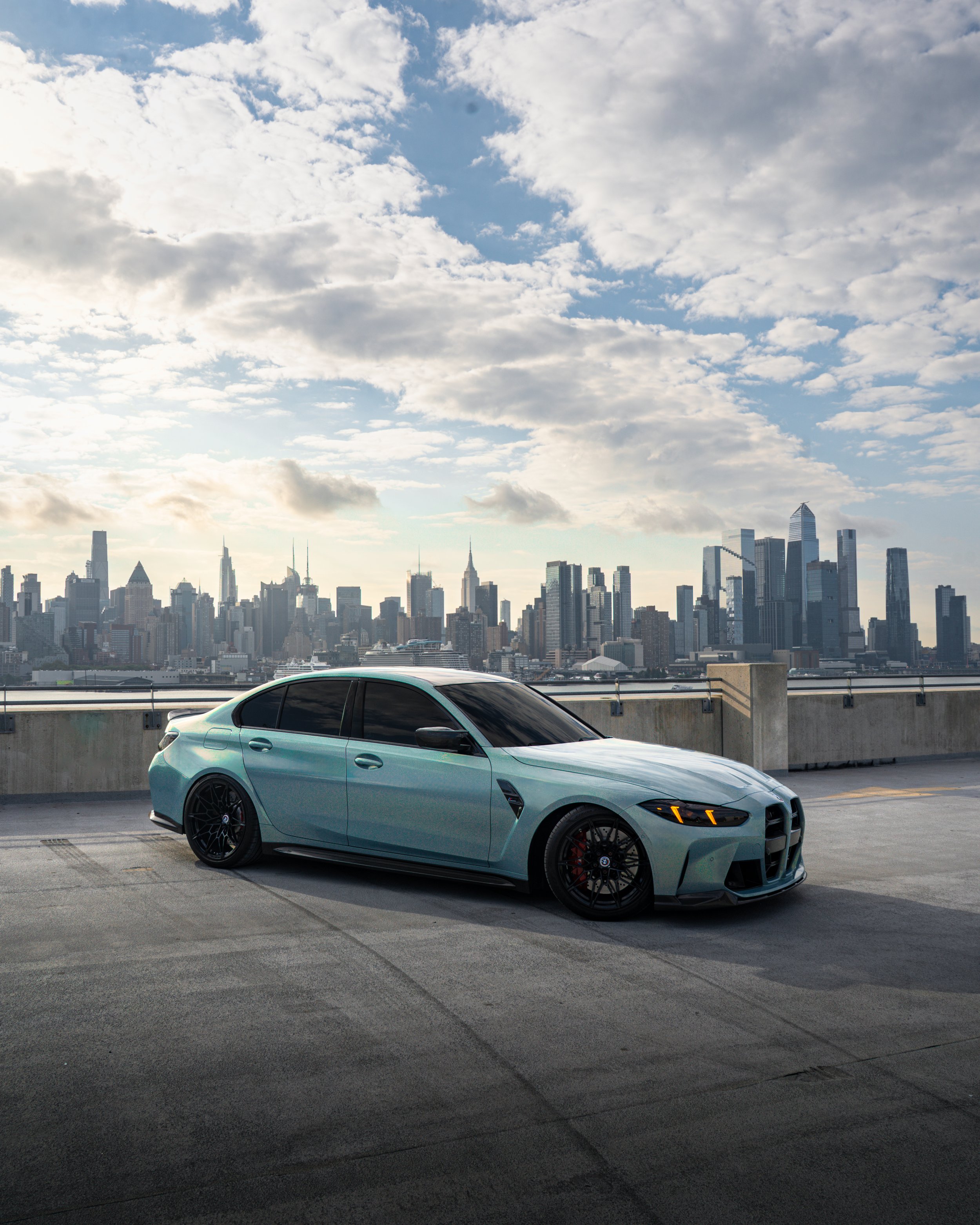 A teal-colored 2025 BMW G80 parked on a rooftop parking lot with the New York City skyline in the background, under a partly cloudy sky.